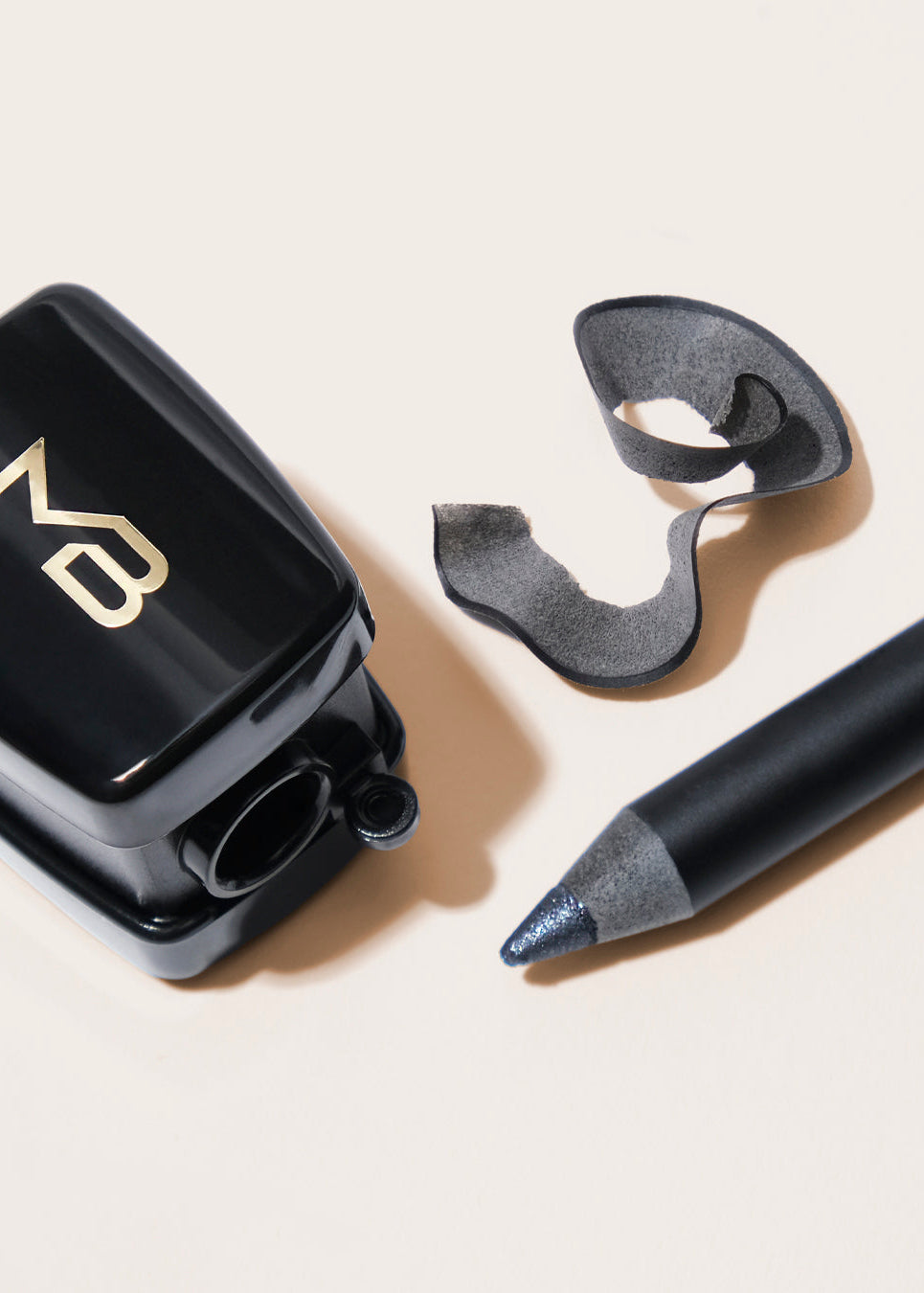 A black cosmetic pencil with a steel gray tip, shown alongside a sharpener with gold lettering and pencil sharpening, all arranged on a light beige background.