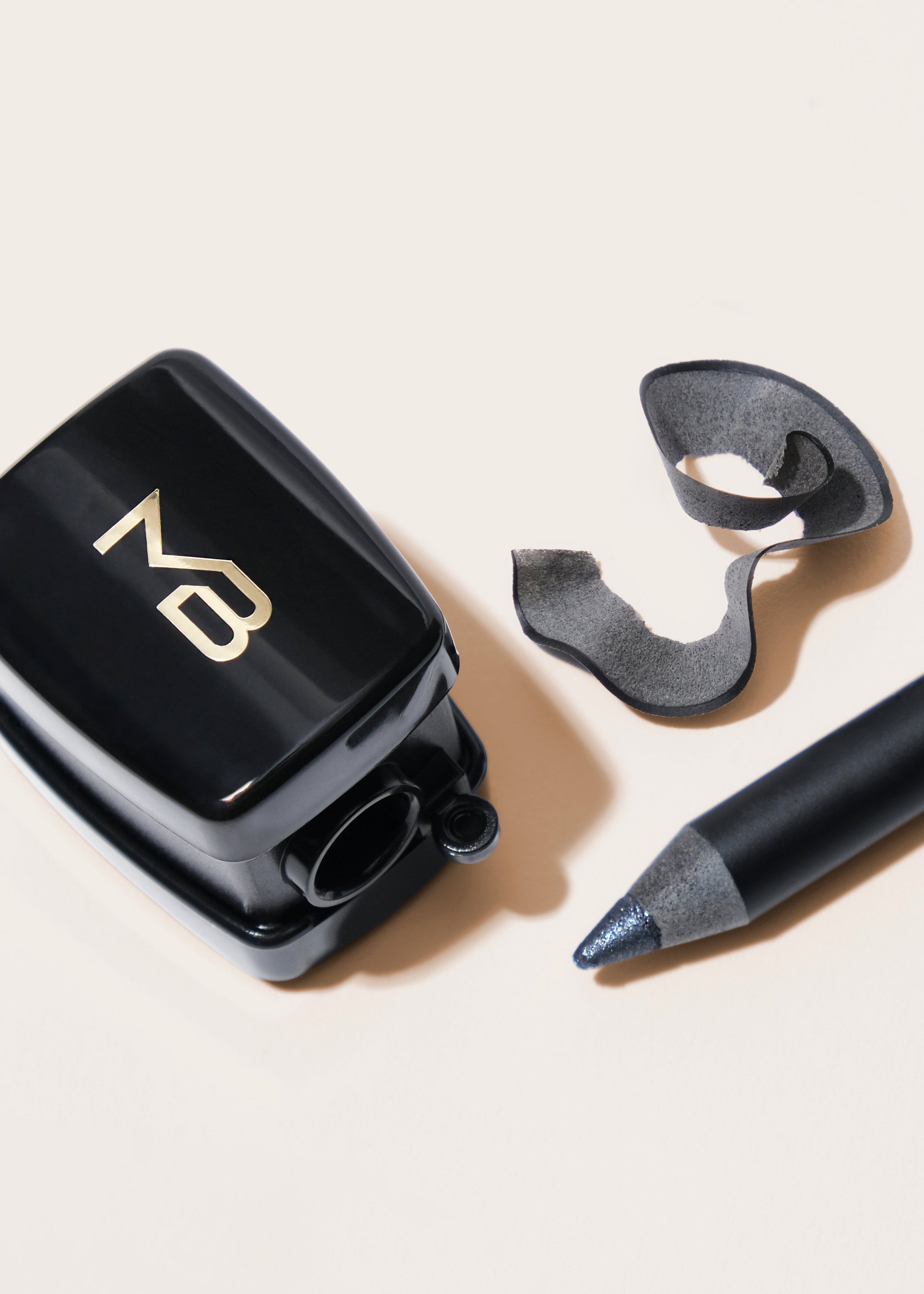 A black cosmetic pencil with gold 'MATURE' branding lies next to a black square cosmetic sharpener with a gold monogram, all arranged on a warm off-white background.