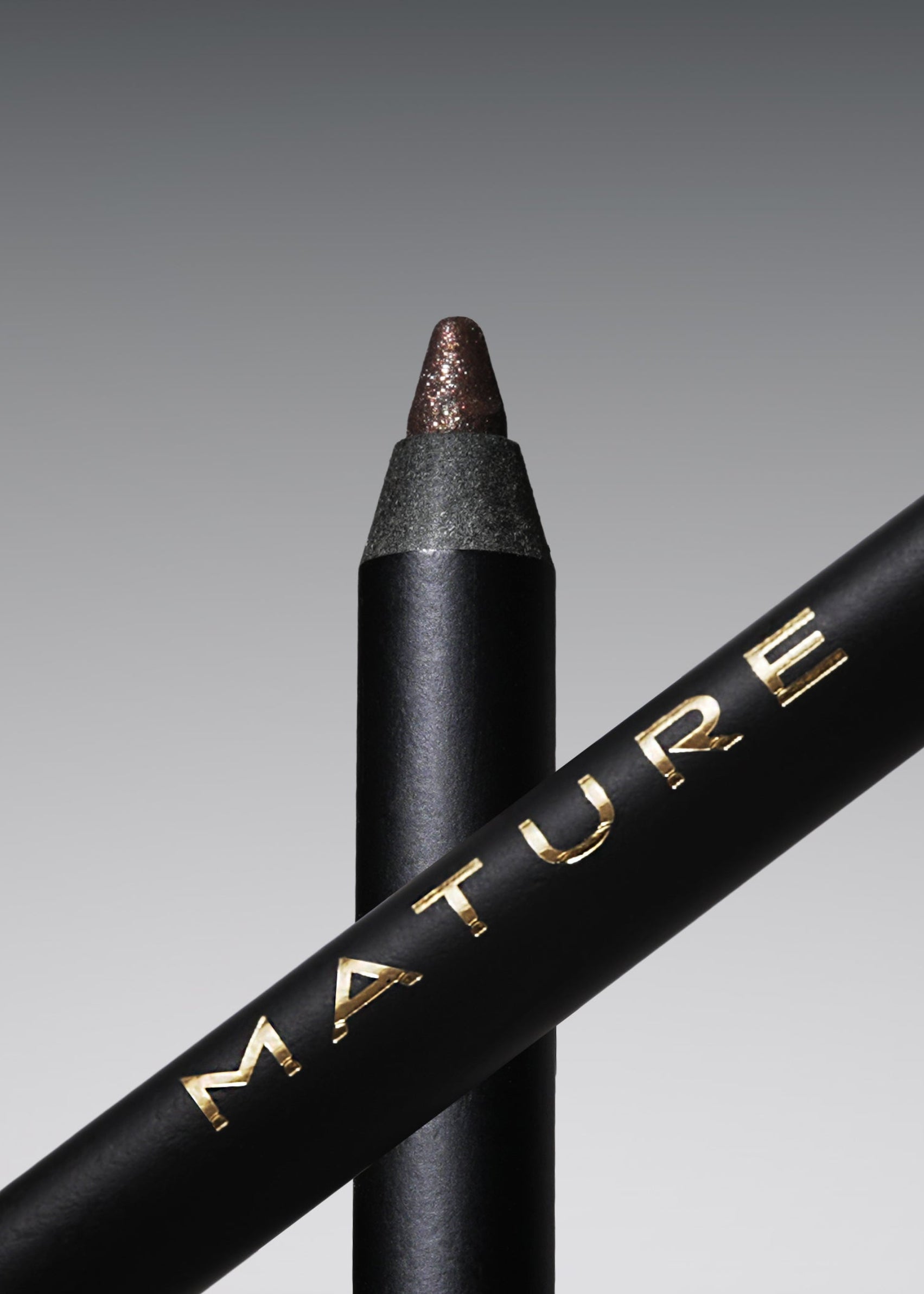 A black cosmetic pencil with gold lettering reading 'MATURE' and a champagne bronze tip, shown alongside a matching black pencil on a gray gradient background.